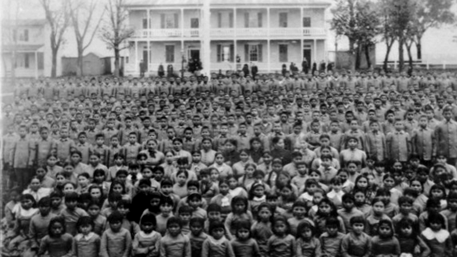 Thousands of students seated in front of a white, two-story school house