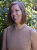 smiling woman with chin-length hair wearing a taupe shirt standing in front of a green bush