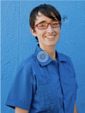 Smiling person with short brown hair, red glasses and a blue shirt in front of a blue wall