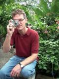 Man in red shirt and jeans taking a photo against a backdrop of green foliage