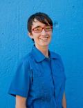 Person with short dark hair wearing red framed glasses and a blue shirt in front of a blue wall