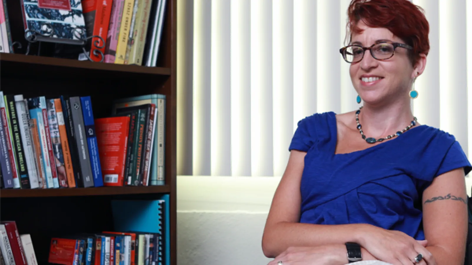 Smiling woman with short dark hair wearing a blue shirt and glasses seated indoors near a bookcase