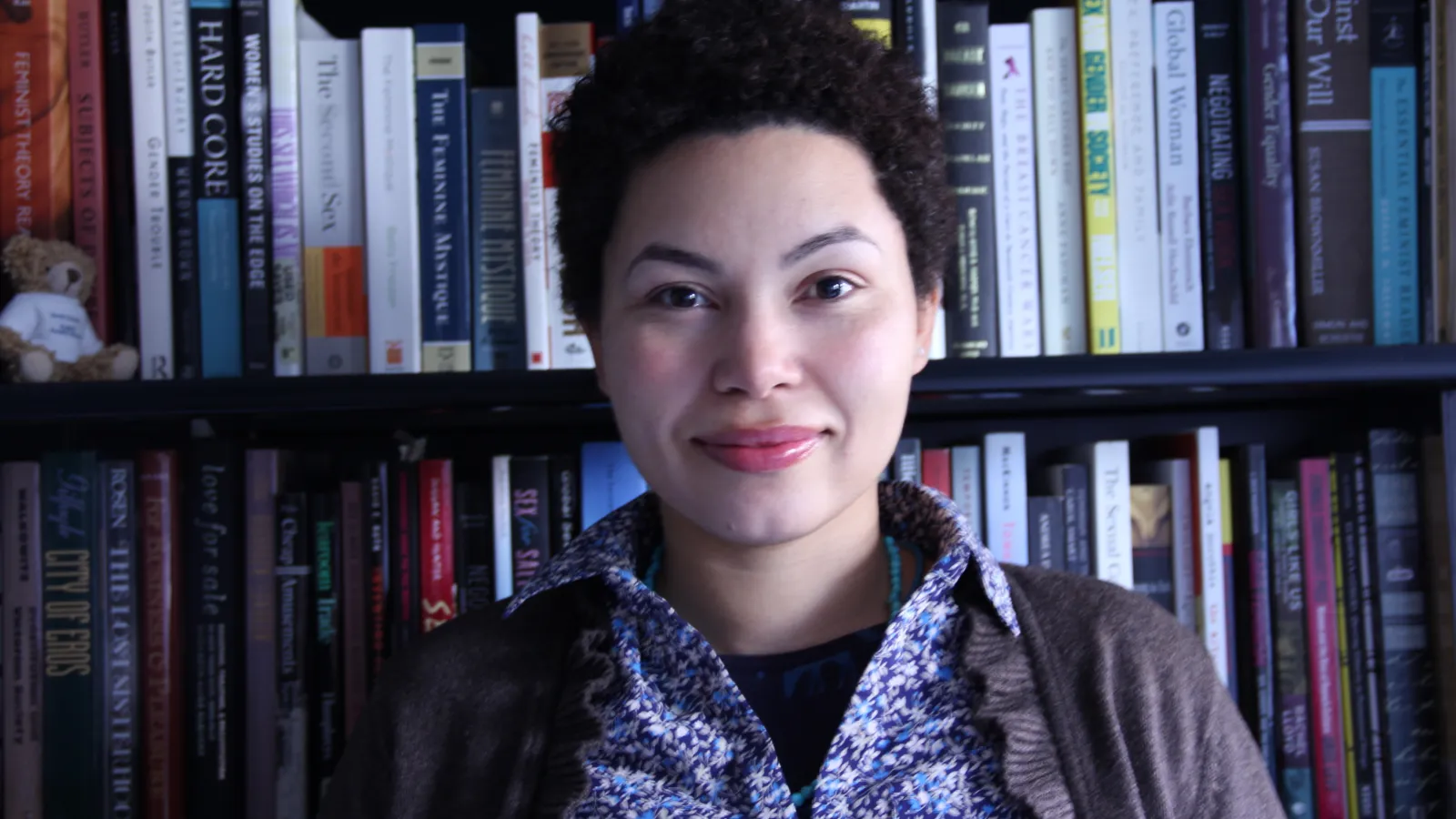 Woman with short dark hair wearing floral shirt and cardigan standing in front of colorful bookcase