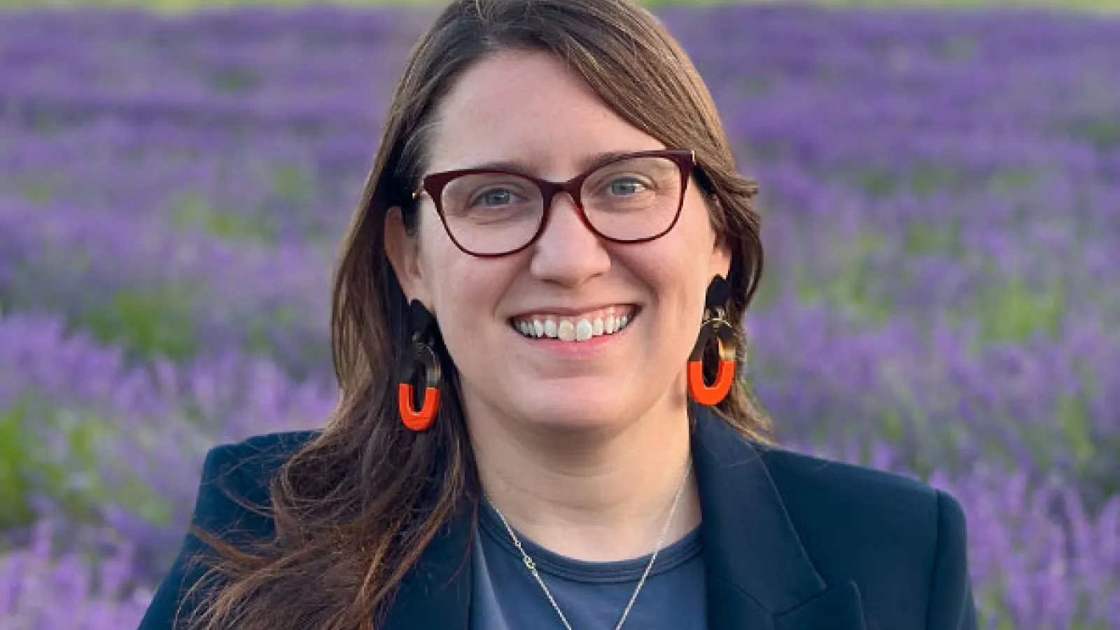 Smiling woman with long hair and glasses standing in field of purple flowers