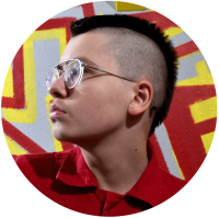Profile photograph of person with a mohawk wearing glasses and a red polo against a graffiti wall