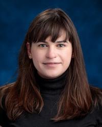 Smiling woman with long dark hair wearing a black turtleneck and posing in front of a blue backdrop