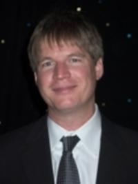 Smiling man with short blonde hair wearing a black suit and tie