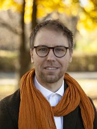 Smiling man with glasses and short hair wearing a black blazer, white shite and orange scarf standing outside in autumn