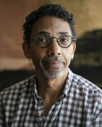 Photograph of a man with short curly hair and a goatee wearing glasses and a gray and white gingham shirt