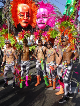 Six male members of Yuma Mas Band dressed in black and white pants with colorful boots ranging in green, pink and orange and matching large, feathered headdresses.