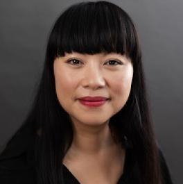 Photograph of woman with long black hair, thick bangs and a dark shirt against a gray background