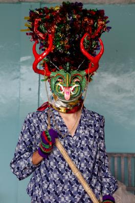 Person wearing a long blue blouse and a green, red and black mask over their face standing in front of a blue backdrop