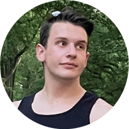 Smiling person with short dark hair wearing a black tank standing in front of a tree