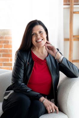 a woman in a grey blazer and red shirt sits on a couch smiling