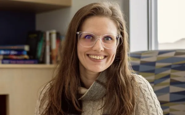 A smiling woman with hands clasped, wearing clear glasses and a tan cardigan