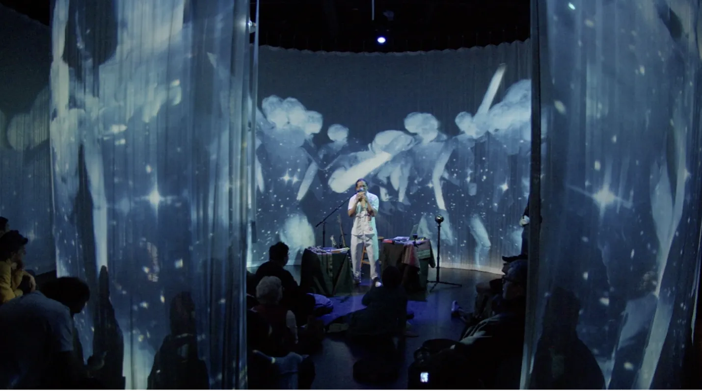 Ati Cachimuel stands at a microphone in the center of a circle of audience members, surrounded by images of dancers and cosmos projected onto hanging curtains.