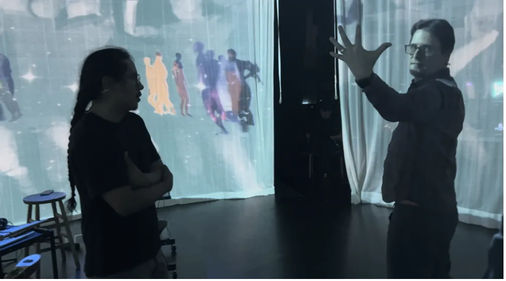 Ati Cachimuel working with Alex Oliszewski (Theater, Film and Media Arts faculty) on stage production for Yana, standing on a stage with projections of dancers on hanging curtains behind them