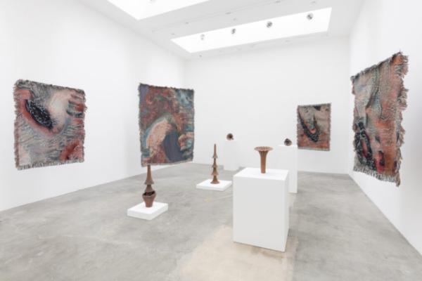 Photograph of gallery with large works on walls and several sculptures on white. pedestals in center