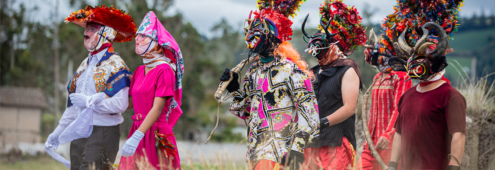 Six people walking through a field of tall grass waring bright costumes and masks covering their faces