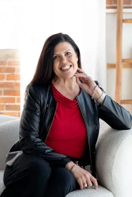 a woman in a grey blazer and red shirt sits on a couch smiling