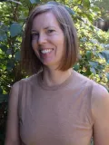 smiling woman with chin-length hair wearing a taupe shirt standing in front of a green bush